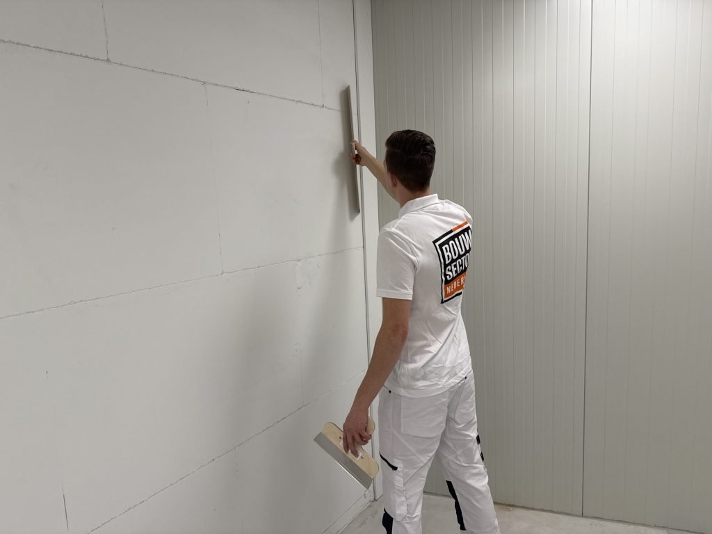 Stukadoor van Bouwsector Nederland in wit werkpak houdt een lange spaan tegen de wand en een plamuurmes in zijn andere hand