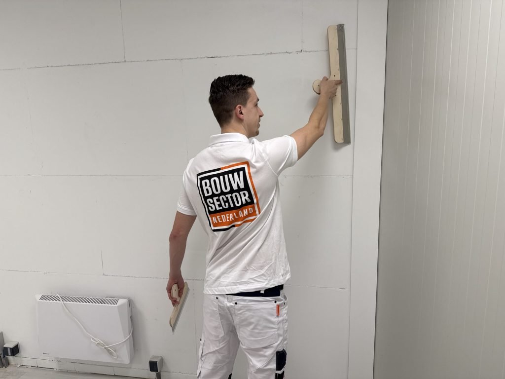 Stukadoor van Bouwsector Nederland houdt een spaan boven schouderhoogte tegen een witte wand