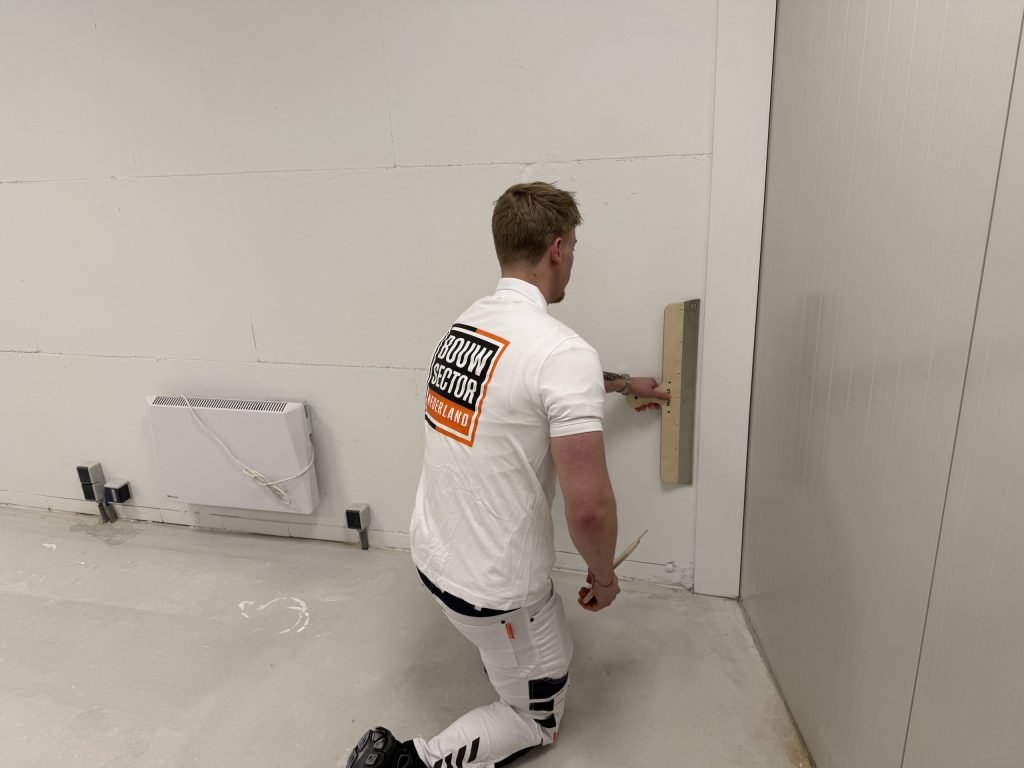 Knielende stukadoor van Bouwsector Nederland zet een lange spaan onderaan tegen een wand naast een radiator