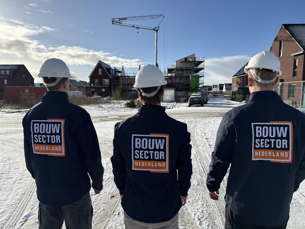 Drie medewerkers van Bouwsector Nederland met witte helm en bedrijfsjas op een besneeuwde bouwplaats met een kraan